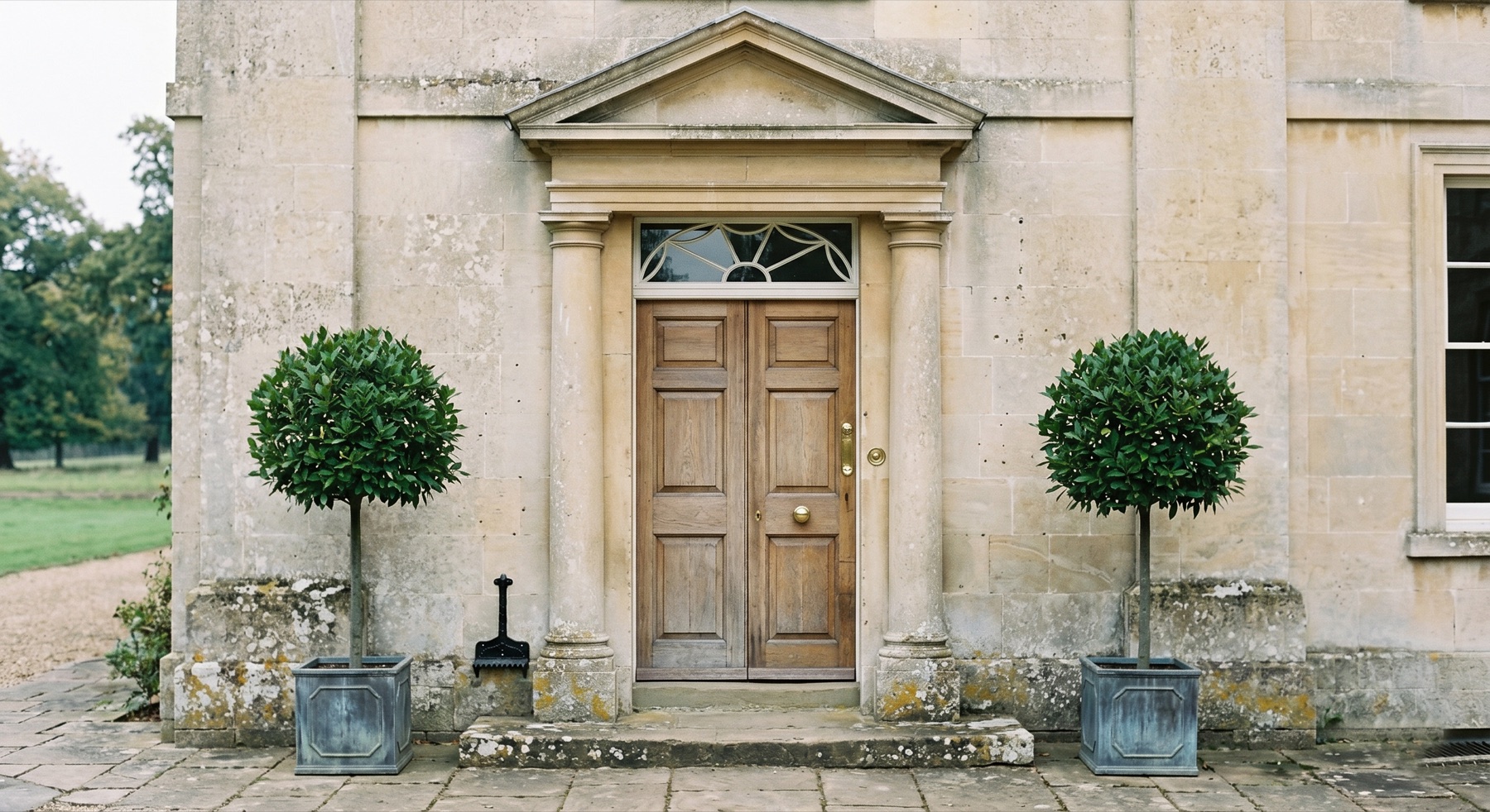 Close view of the pedimented front door in Bath limestone, flanked by two clipped bay topiary in lead planters.